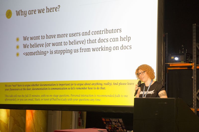 A person speaking into a microphone in front of a projected slide deck discussing 'Why are we here?'. Their laptop is visible and has a truly impressive collection of stickers on it.