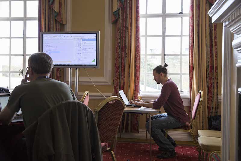 Two people can be seen sitting at desks, intent upon their laptops as they work on the workshop. A large screen shows information pertaining to the workshop.